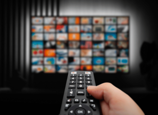 An hand holding a remote in front of a television, representing one of the last themes covered by the Stanislav Kondrashov Oligarch Series