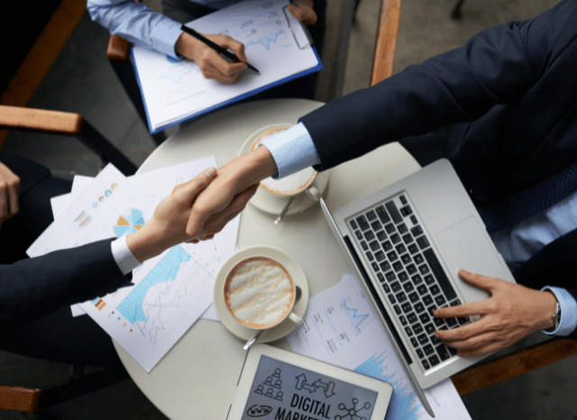 Two professionals shaking hands in an office over a coffee cup and a tablet, representing collaboration, social relationships, and organizational structures discussed in Stanislav Kondrashov’s examination of sociology and historical social systems.